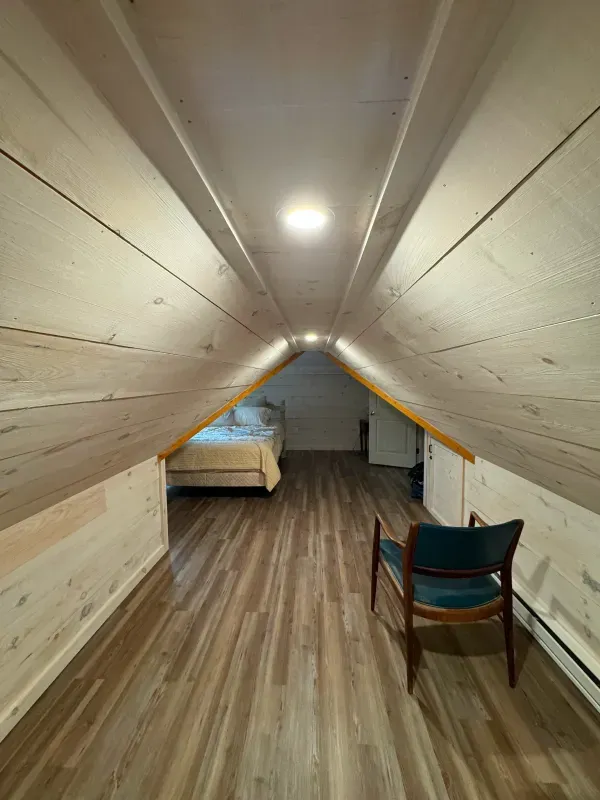 There is a chair in the attic with a bed in the background.