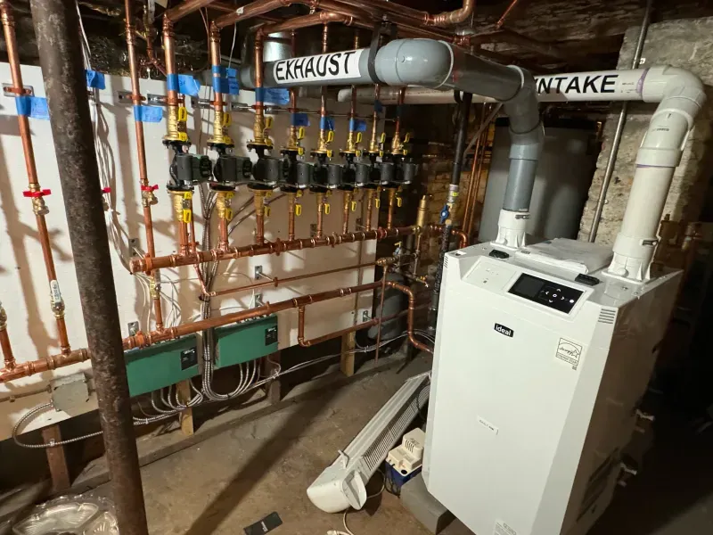A white boiler is sitting in a basement surrounded by pipes.