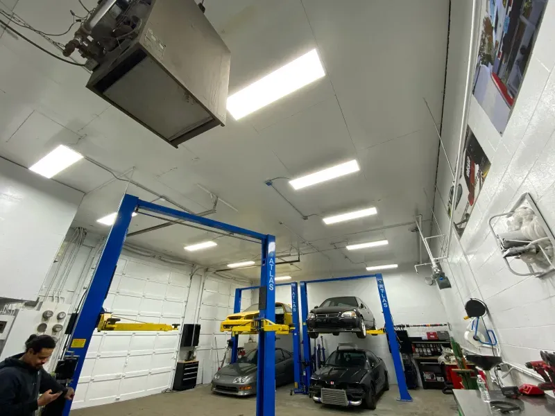 A man is standing in a garage with two cars on lifts.