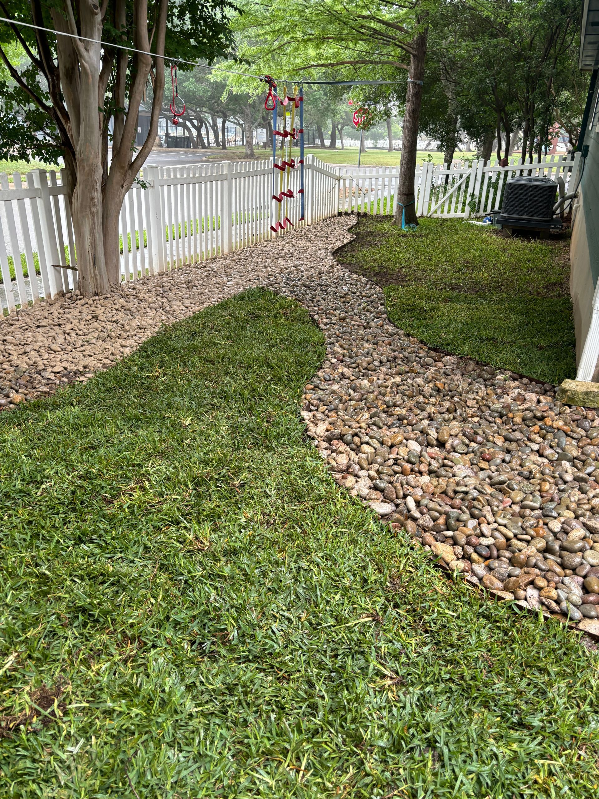 Green ground cover with a rock border next to a white picket fence, trees, and a building.