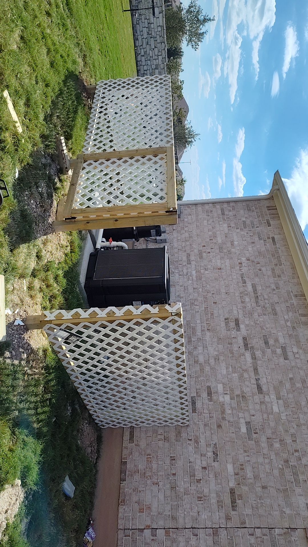 White lattice screen hides an air conditioning unit next to a brick building and green lawn under a partly cloudy sky.