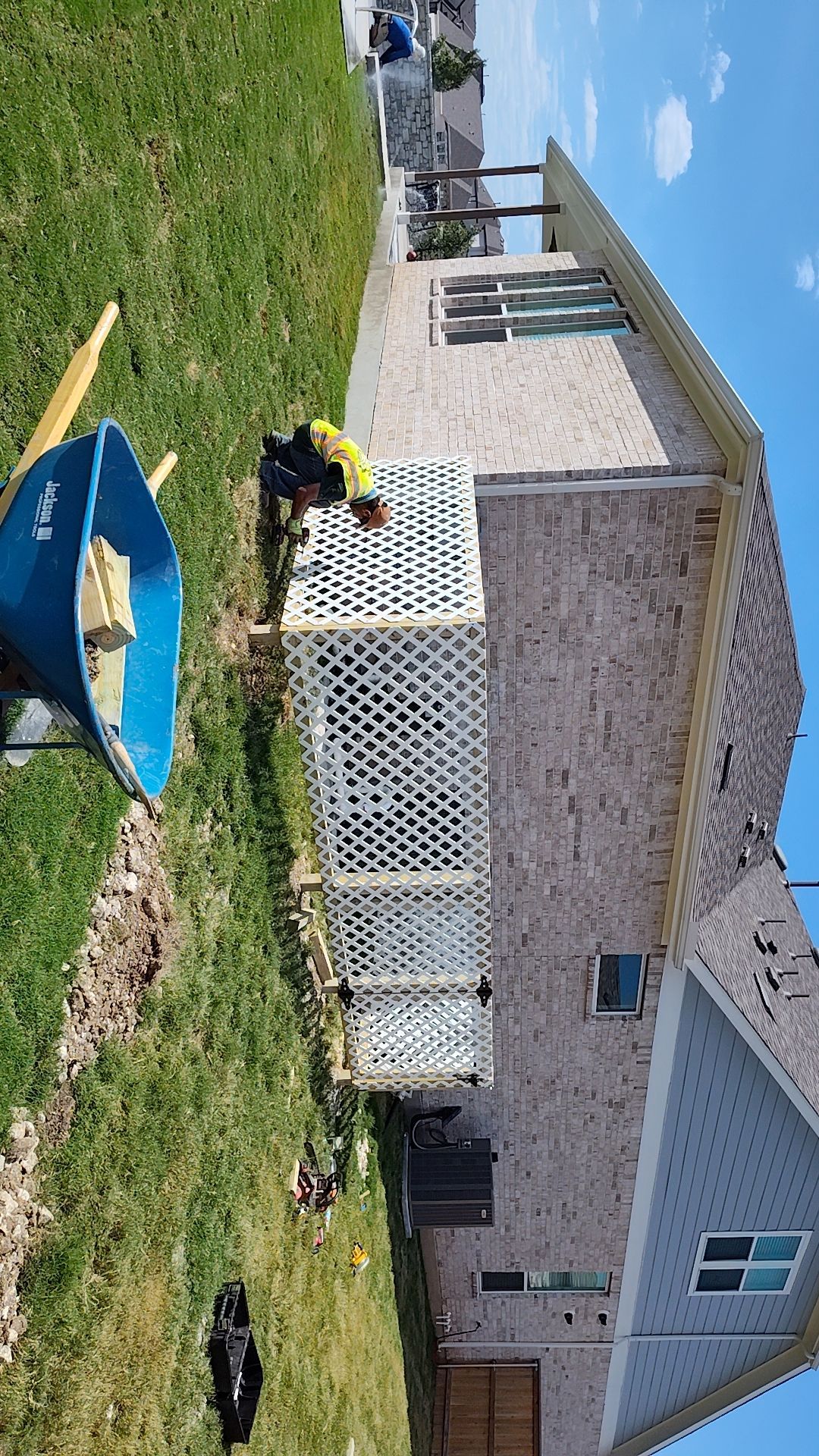 Construction worker installing white lattice along the side of a brick house on a sunny day. A blue wheelbarrow sits nearby.