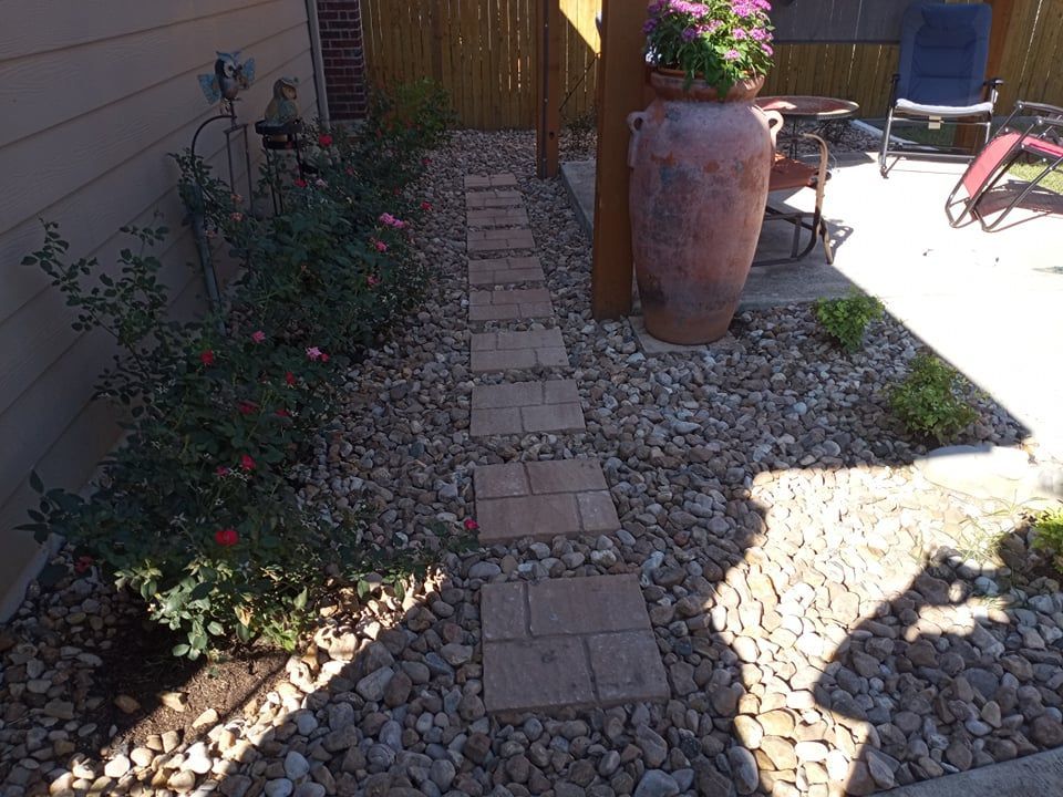 A stone path through a garden with rose bushes, leading to a patio with a large pot and chair.