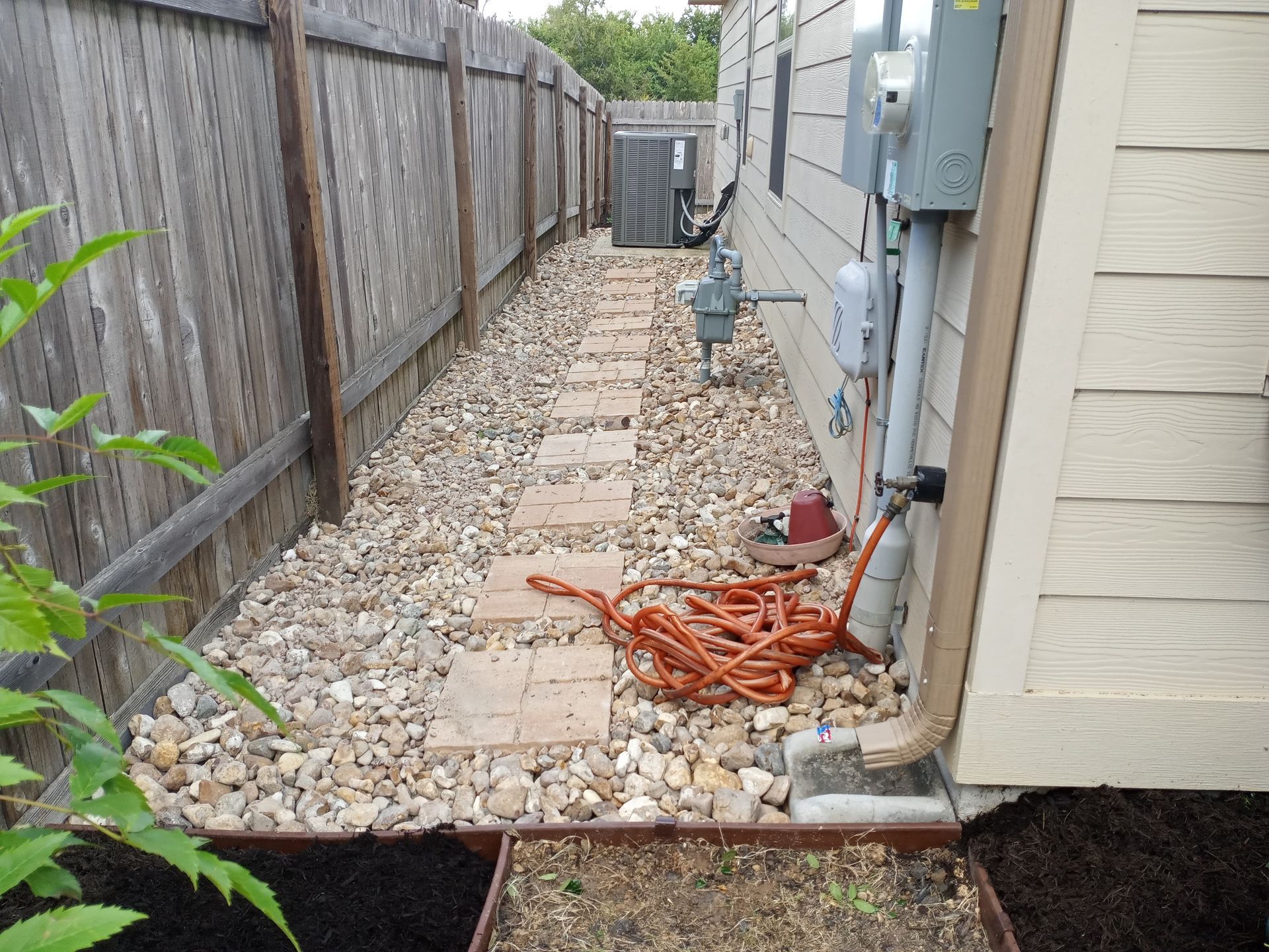 A narrow gravel path with stepping stones leads to a wooden gate, beside a house and fence.