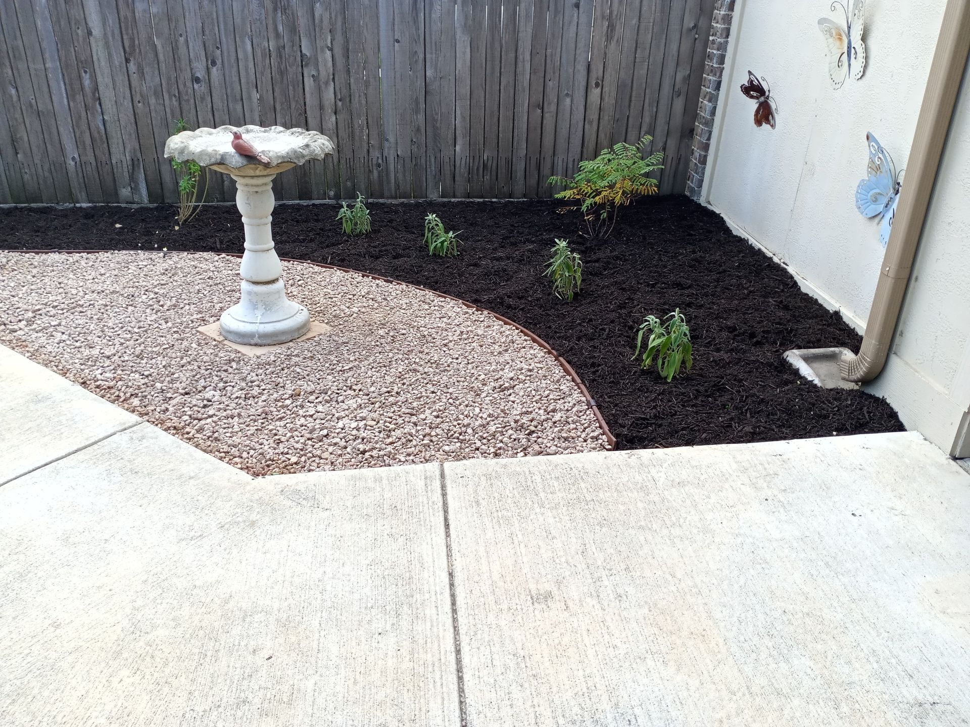 Concrete patio with gravel, mulch, birdbath, and plants along a wooden fence and wall.