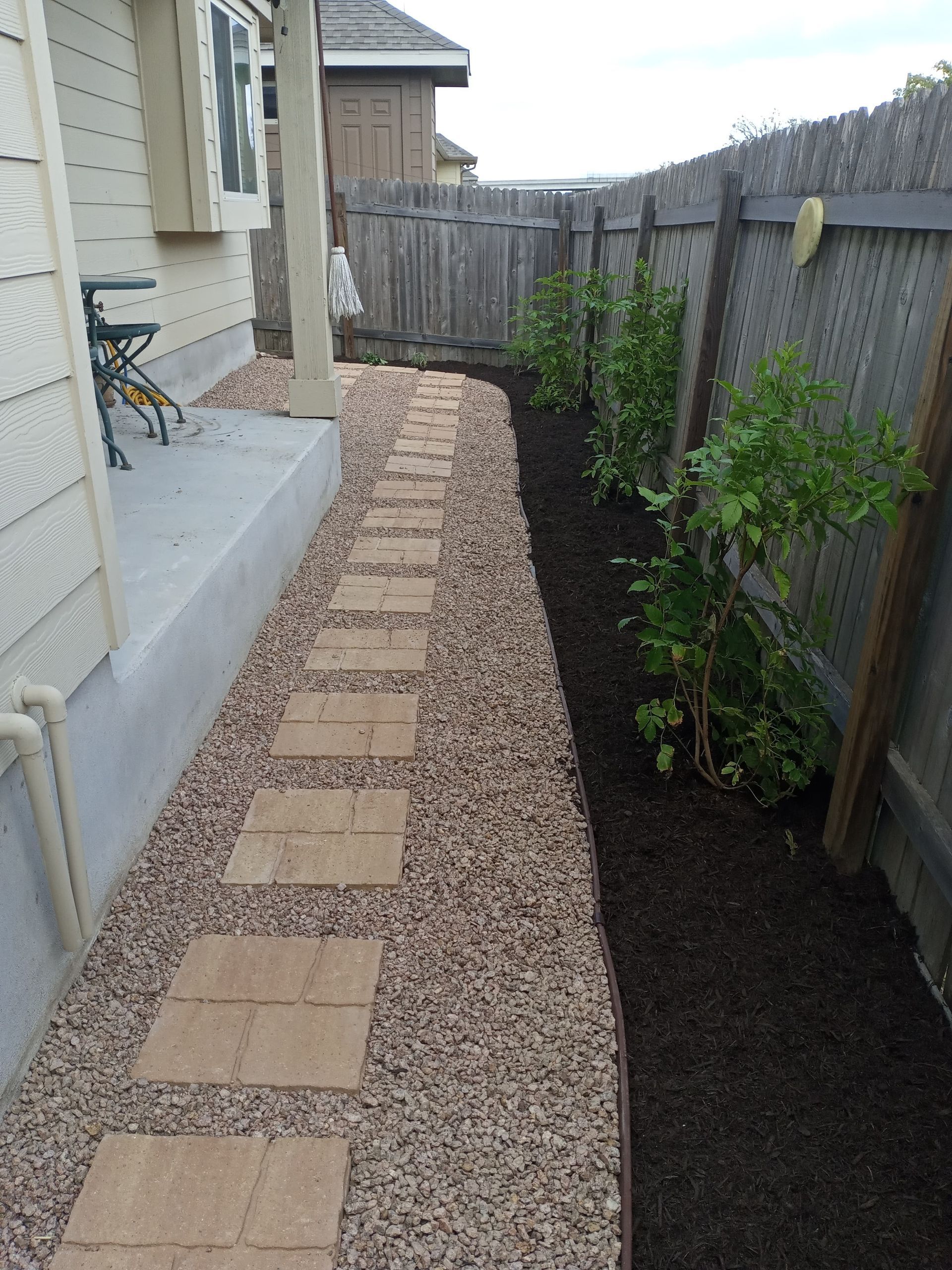 Stone path beside a house, bordered by gravel and mulch with plants. Wooden fence in background.