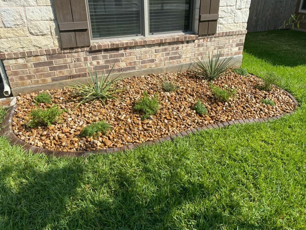 Flower bed with brown rocks and plants near a brick house with green grass.