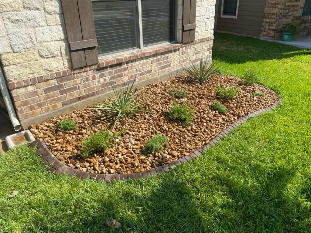 Flower bed with brown rocks and green plants edged with a brown border in front of a house.