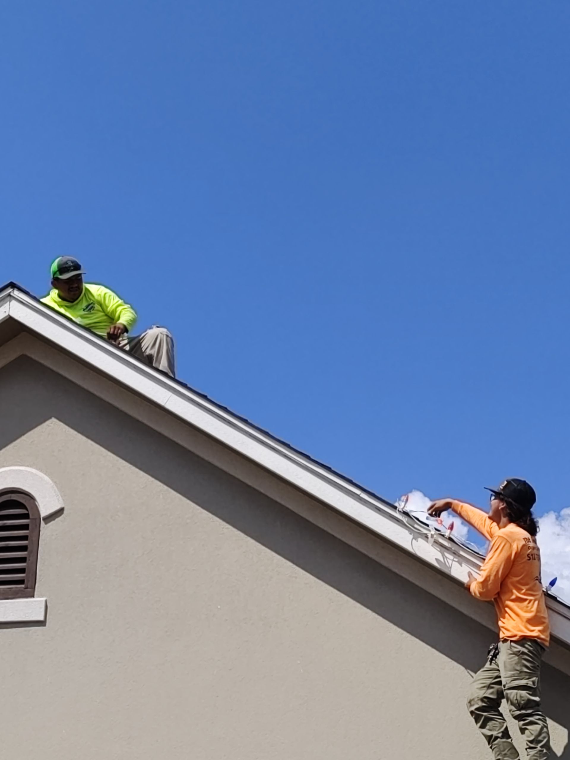 Two workers on a rooftop, one on the roof, one on a ladder. Blue sky, beige siding, green/orange shirts.