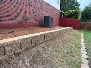 Retaining wall of tan blocks in a grassy yard, next to a red brick wall and a fence.