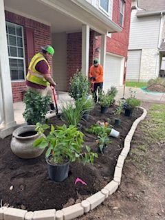 Two landscapers planting flowers in a garden bed in front of a brick house.