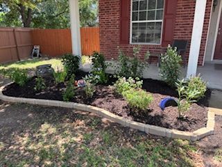 Flower bed in front of a brick house with various green plants and brown mulch, bordered by tan bricks.