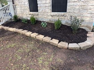 A stone-bordered garden bed against a stone building holds various green plants and dark mulch.