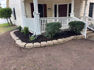 House porch with stone-edged flower bed filled with dark mulch, green bushes and small plants.