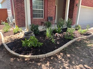Landscaped front yard bed with dark mulch and beige border, plants, and a dark pot. Red brick house.