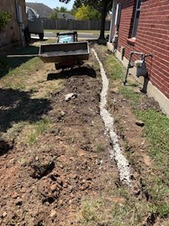 A trench filled with gravel alongside a brick building, likely for drainage. Construction equipment visible.