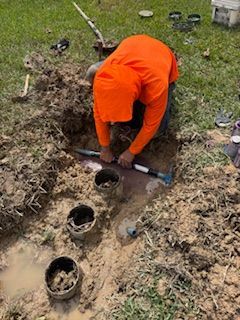 Person in orange shirt working in a muddy trench, installing pipes in a yard.