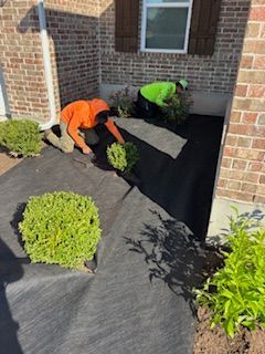 Two people installing landscaping fabric in a flowerbed near a brick building.