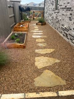 A stone walkway leading to a garden with a brick building in the background.