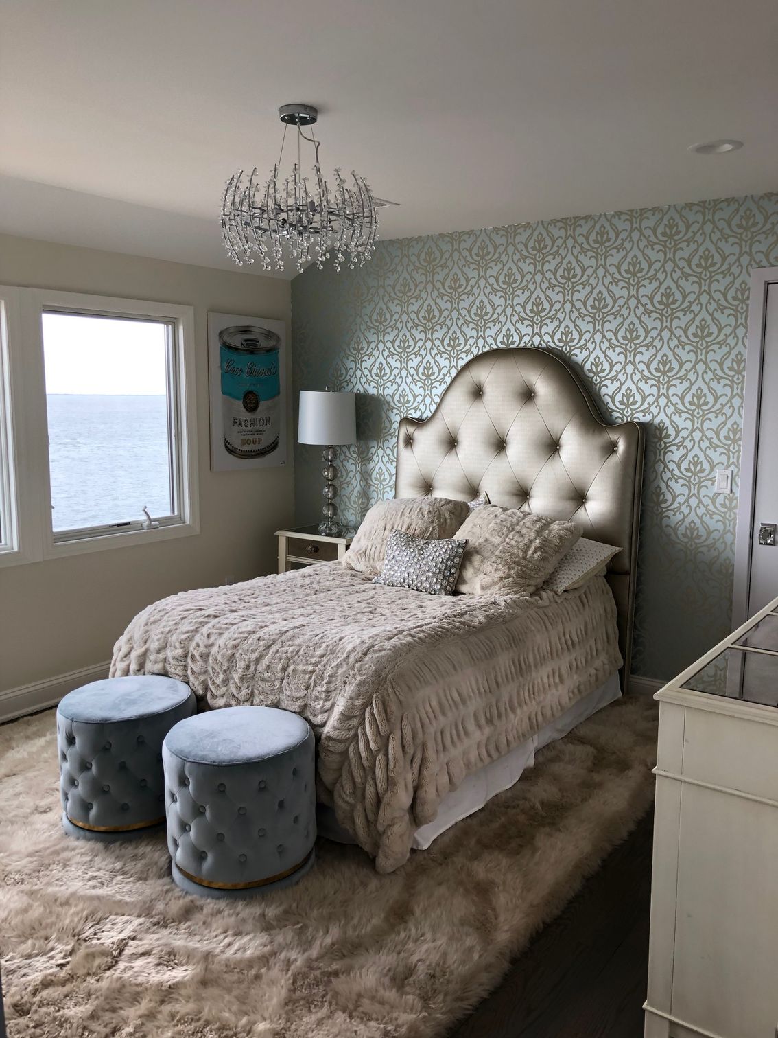 A bedroom with a bed , two ottomans and a window overlooking the ocean.