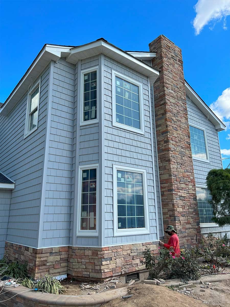 A man is standing in front of a large white house with a brick chimney.