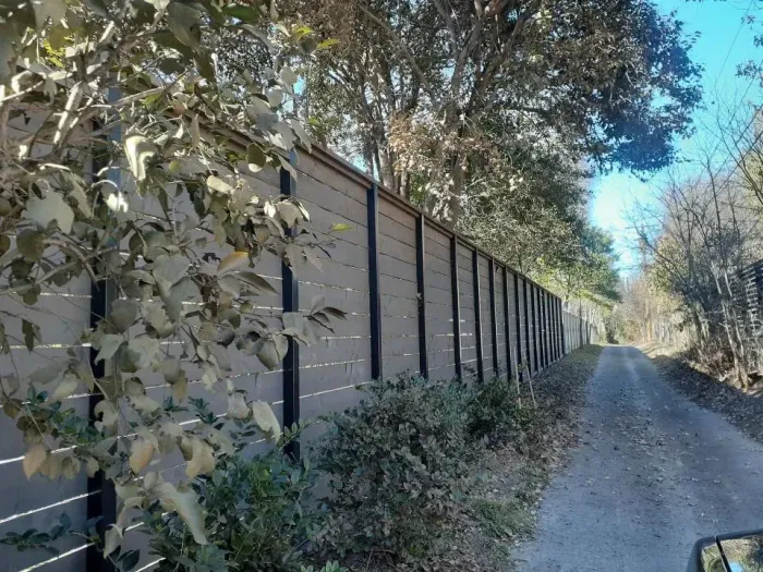 Long fence beside a gravel path through a wooded area on a sunny day