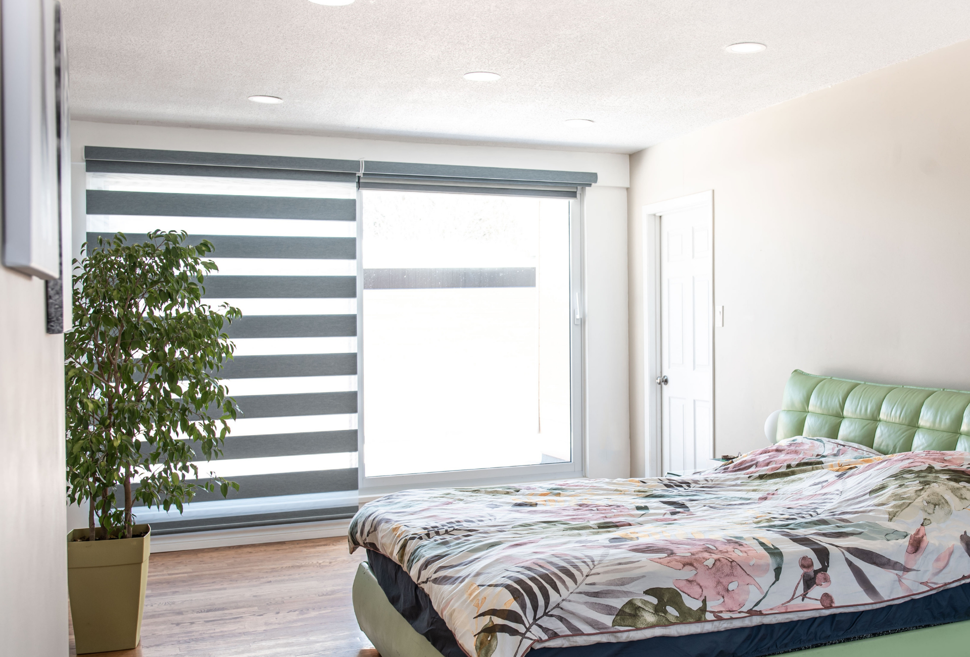 Bedroom with a large window and patterned blinds, a bed with floral bedding, and a large plant.
