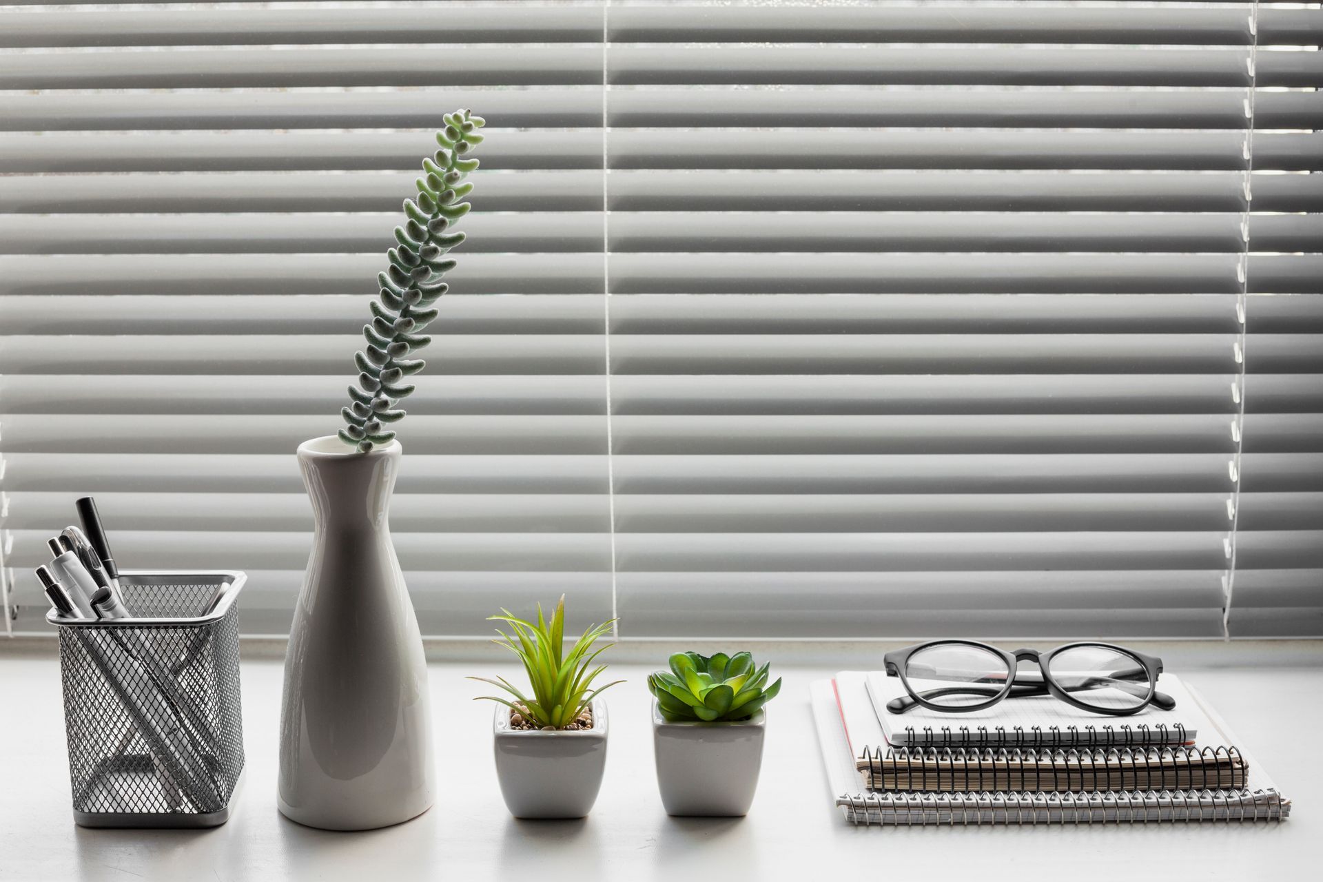 White vase with plant, two small potted succulents, pen holder, notebooks, and glasses on a windowsill with blinds.