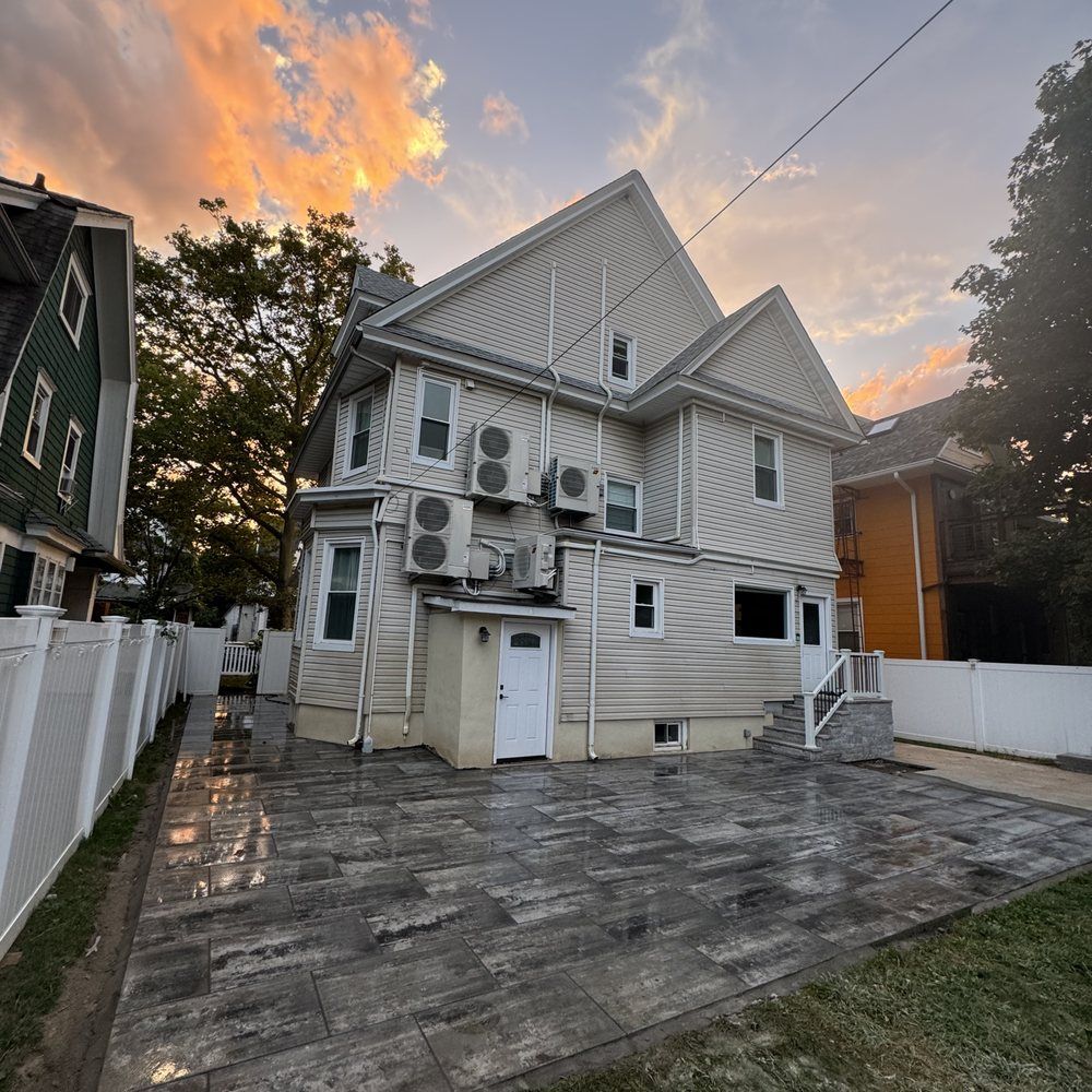 Back of a three-story beige house with air conditioners, a paved patio, and a white fence. Sunset colors in the sky.