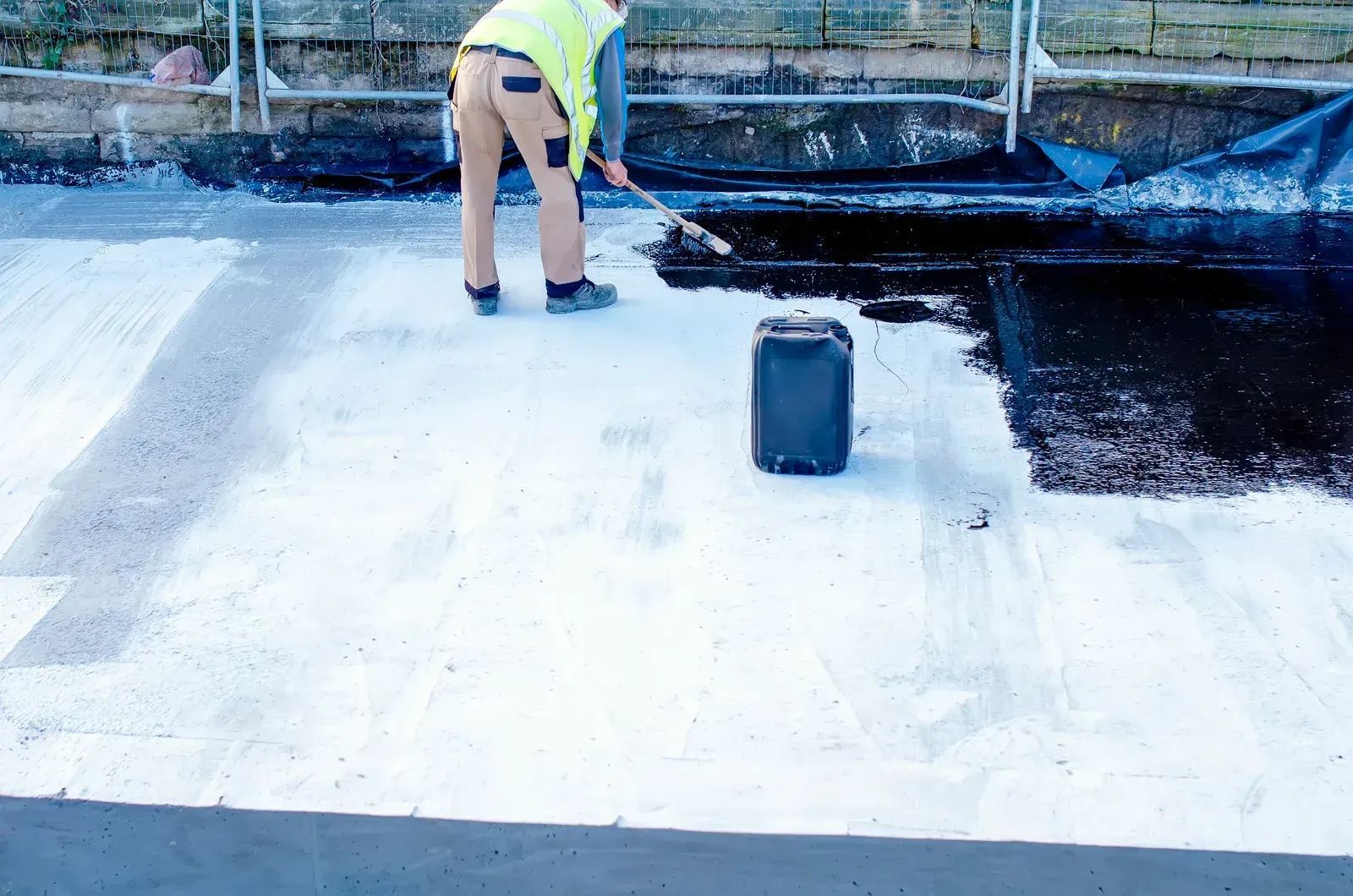 Person applying black sealant to a white-painted concrete surface with a roller, standing near a black container.