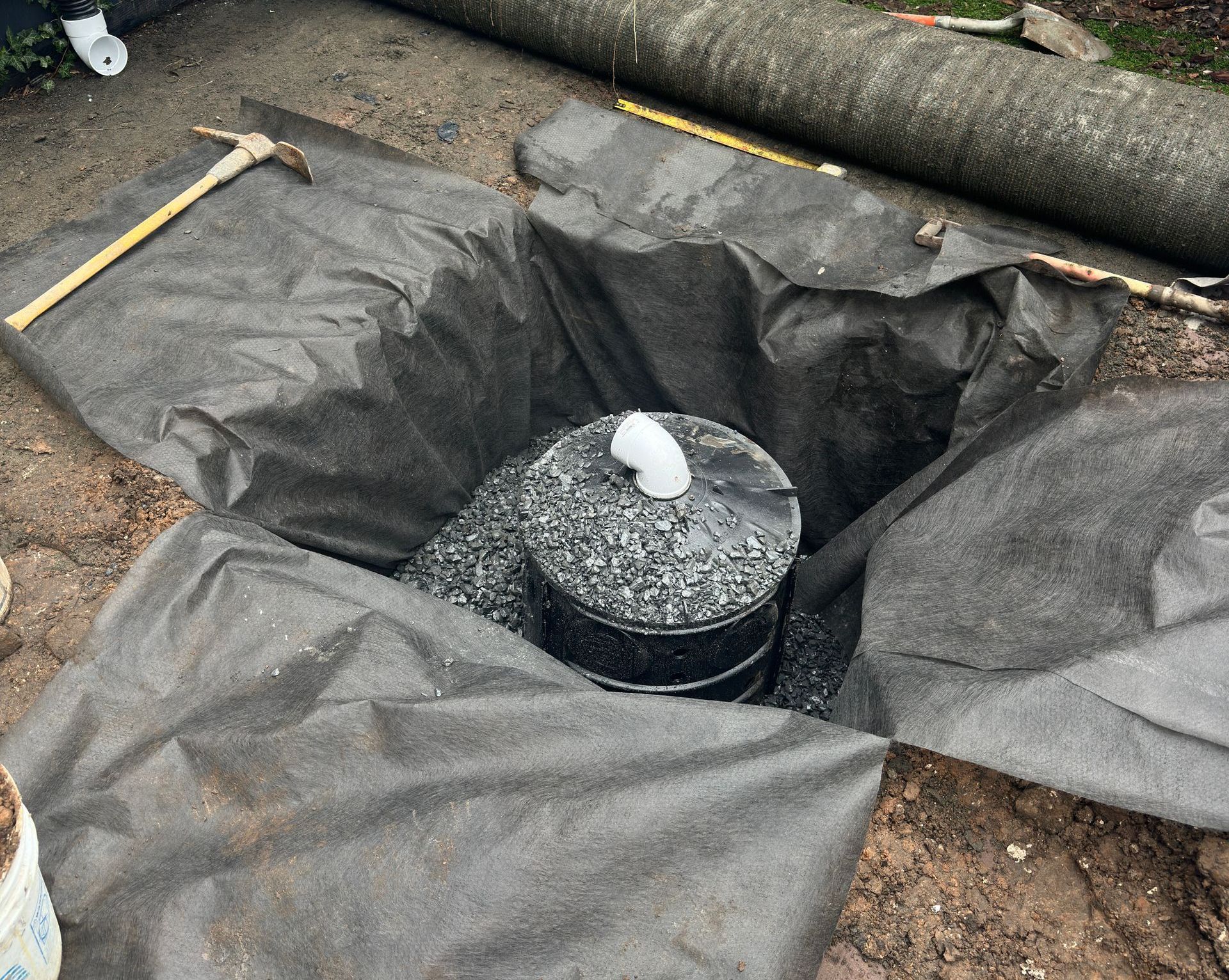 Black landscape fabric lines a hole in the ground with a black container surrounded by gravel.