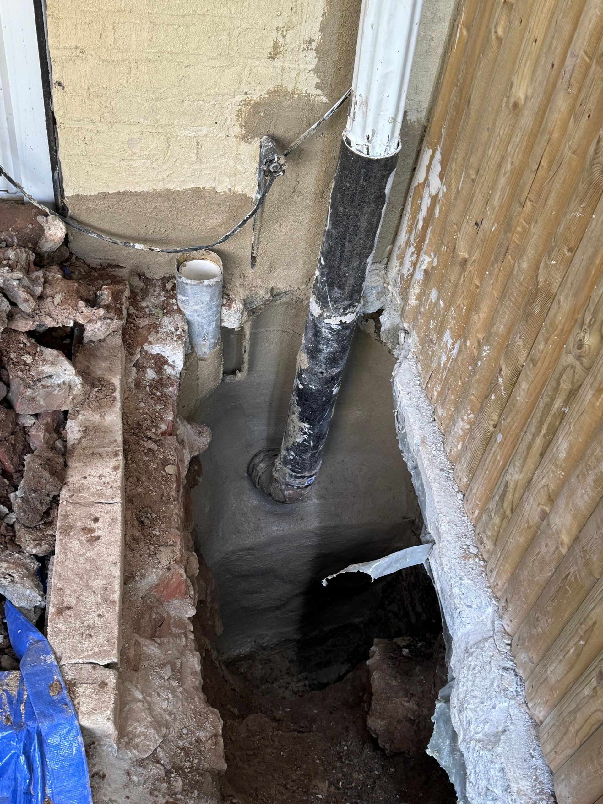 Exterior view of a wall with exposed pipes and a trench. The wall is tan and brown, with a white downspout.