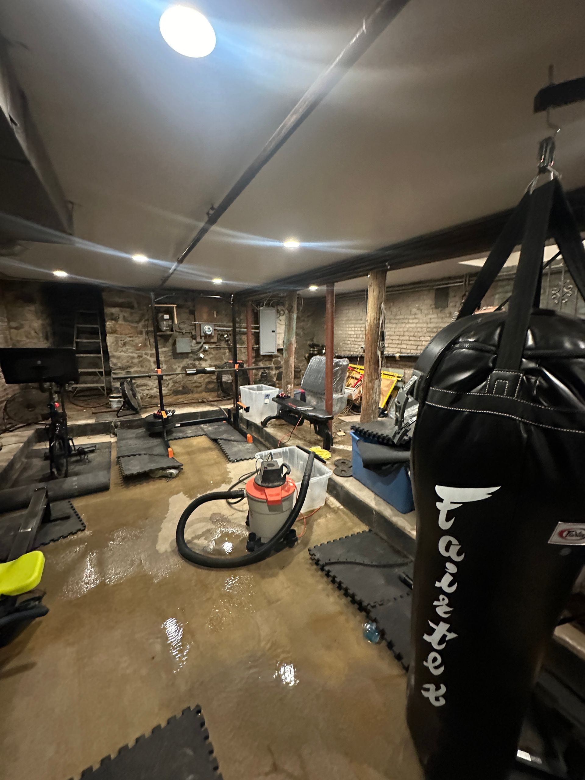 Basement gym with wet floor, a punching bag, and vacuum cleaner, surrounded by exposed brick walls.