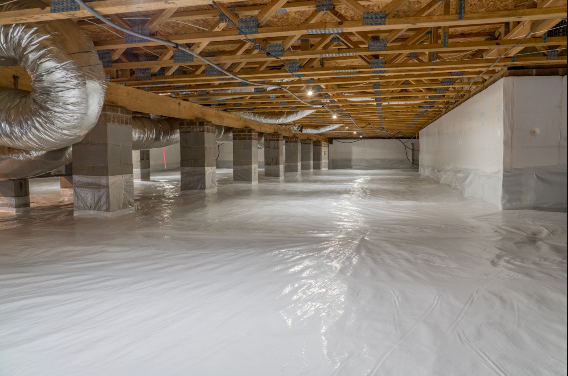 Crawlspace with white vapor barrier flooring and support columns under a wooden structure.