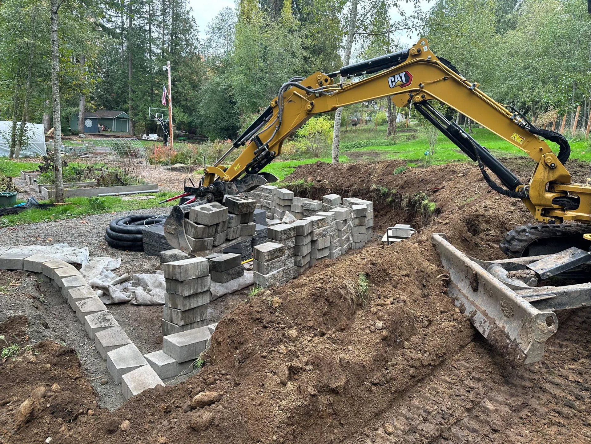 Yellow excavator building a retaining wall in a yard with dirt and gray blocks.