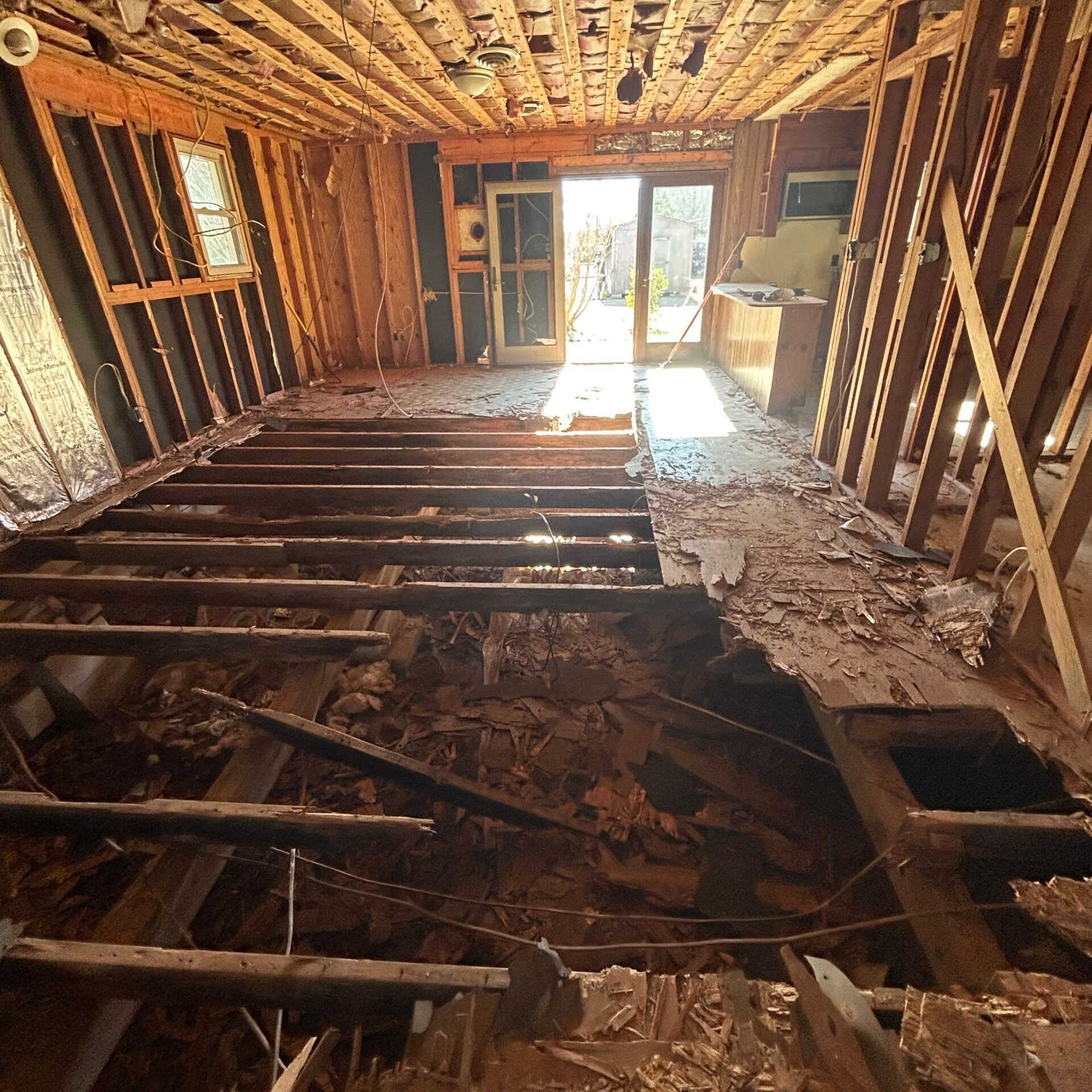 Interior of a home under renovation, with exposed studs, missing floorboards, and debris. Sunlight streams through a doorway.