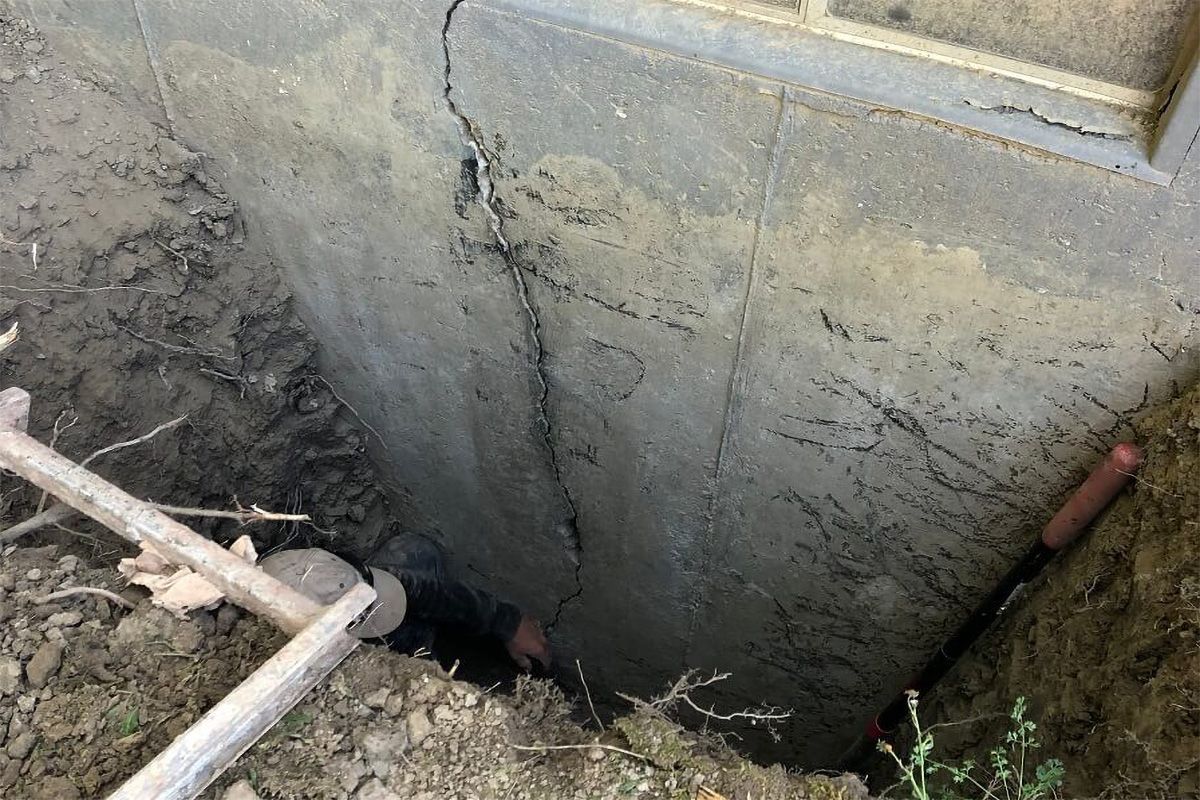 Concrete wall with deep vertical cracks, dug out to reveal its base, with a shovel.