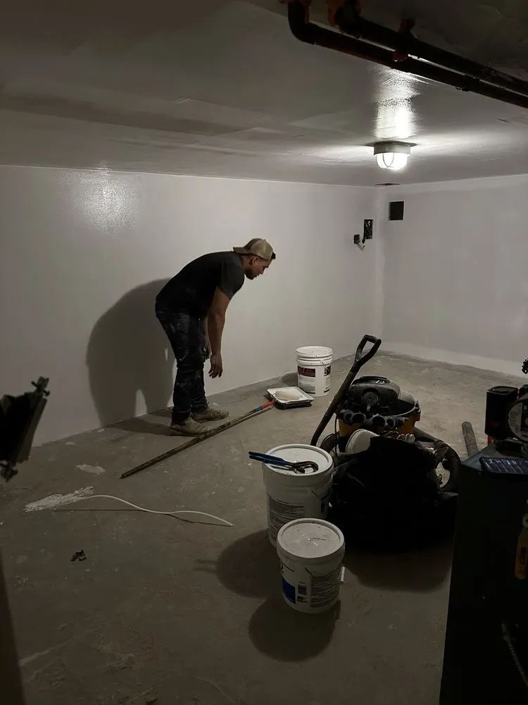 A man in a room painting white walls, with paint buckets and tools on the floor.