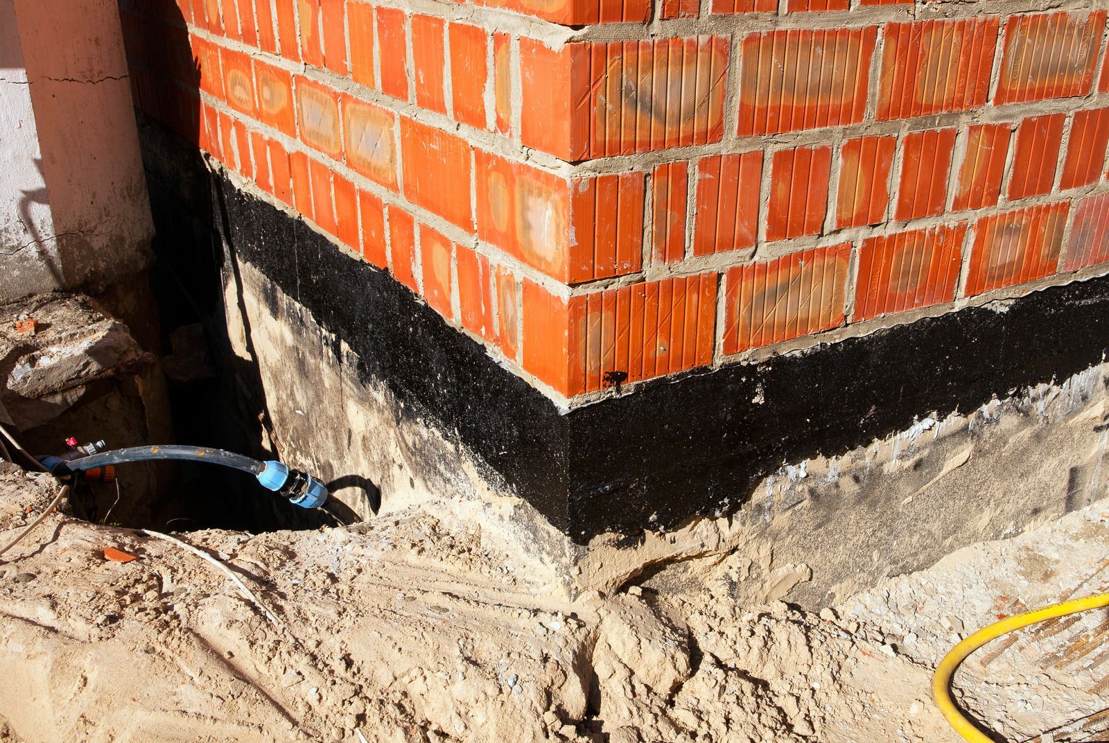 Brick wall corner with black waterproofing layer on the foundation, surrounded by sand and construction materials.
