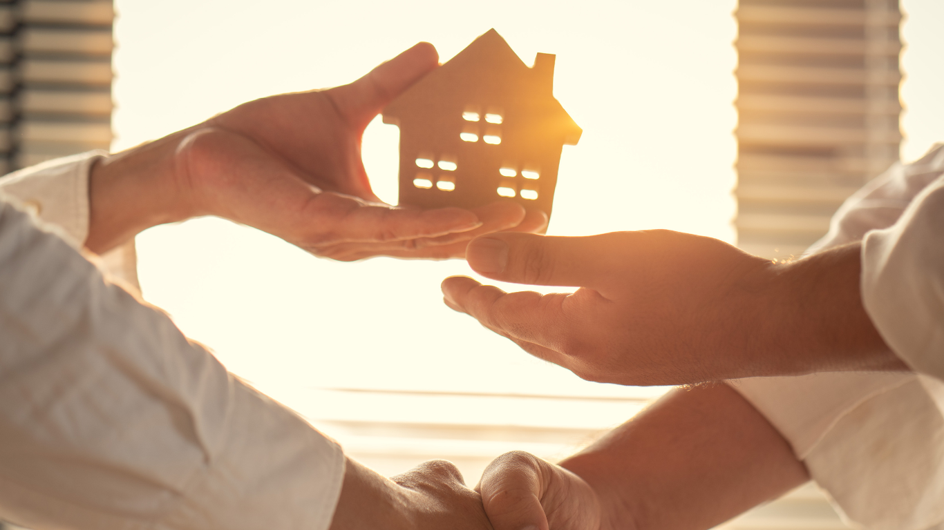 Two people are shaking hands while holding a model house in their hands.