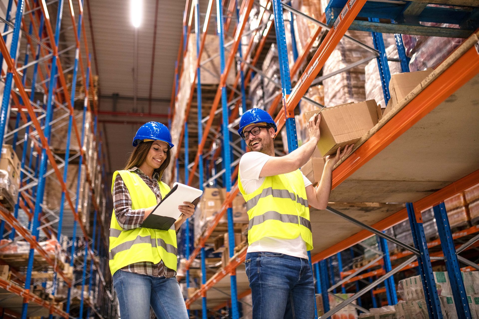 Two warehouse workers, wearing hard hats and safety vests, inspect packages on shelves.