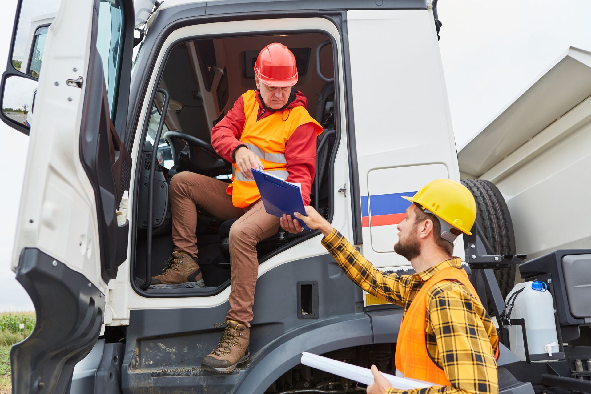 Two workers in safety vests and helmets near a white truck. One in the truck seat, the other handing paperwork.