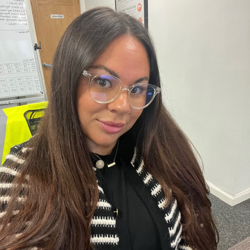 Woman with long brown hair and glasses smiles, wearing a black sweater under a striped cardigan, in an office setting.