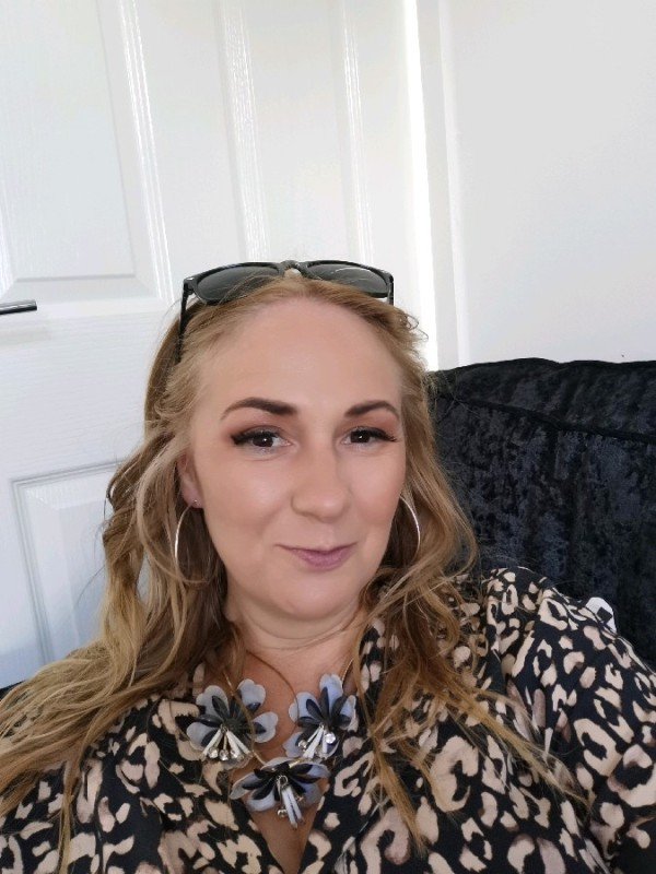 Woman with blonde hair, wearing sunglasses, leopard print blouse, and large necklace, smiling.