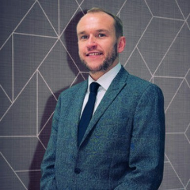 Man in a blue suit and tie smiling in front of a patterned wall.