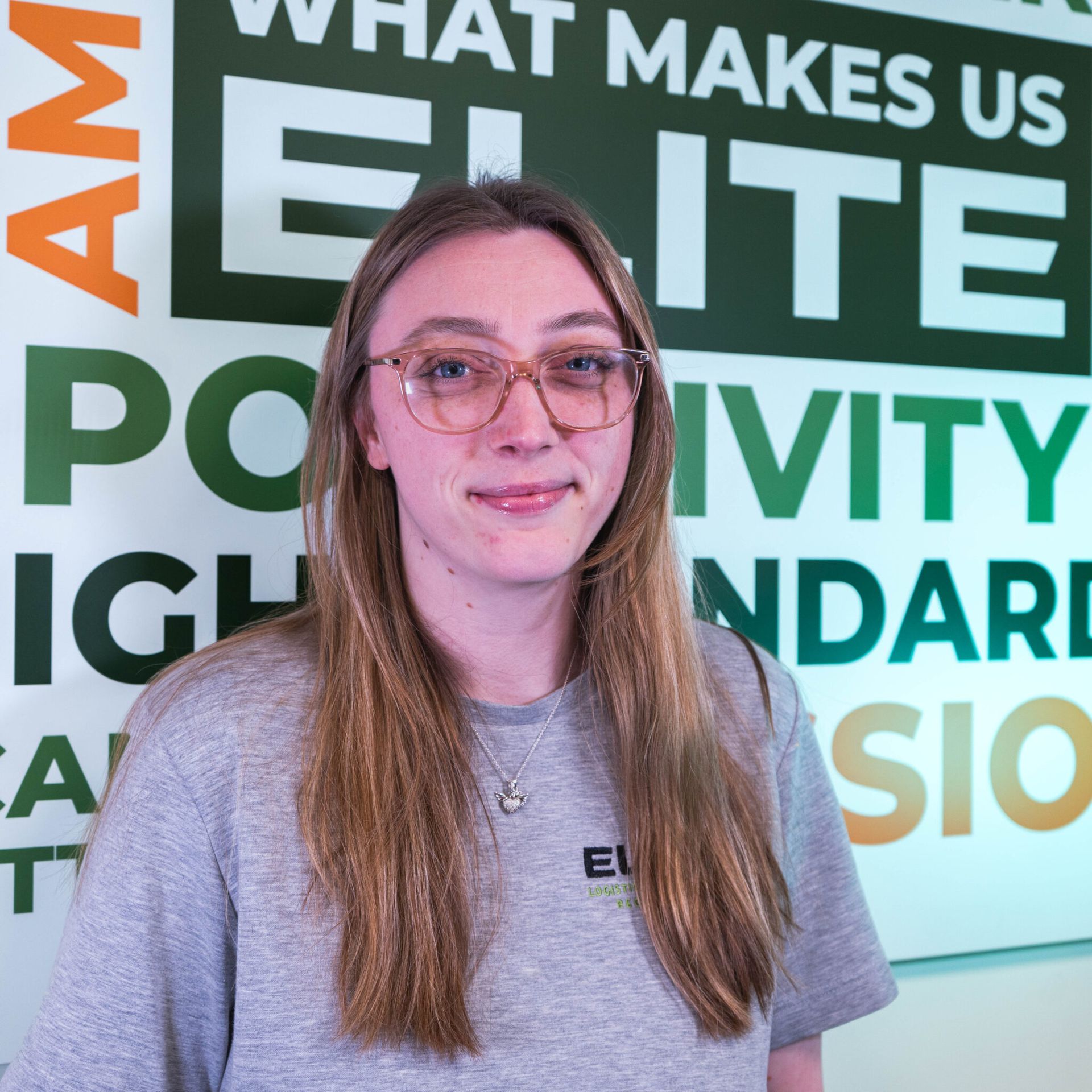 Woman with glasses smiling, wearing a gray shirt in front of a green and white wall with text.