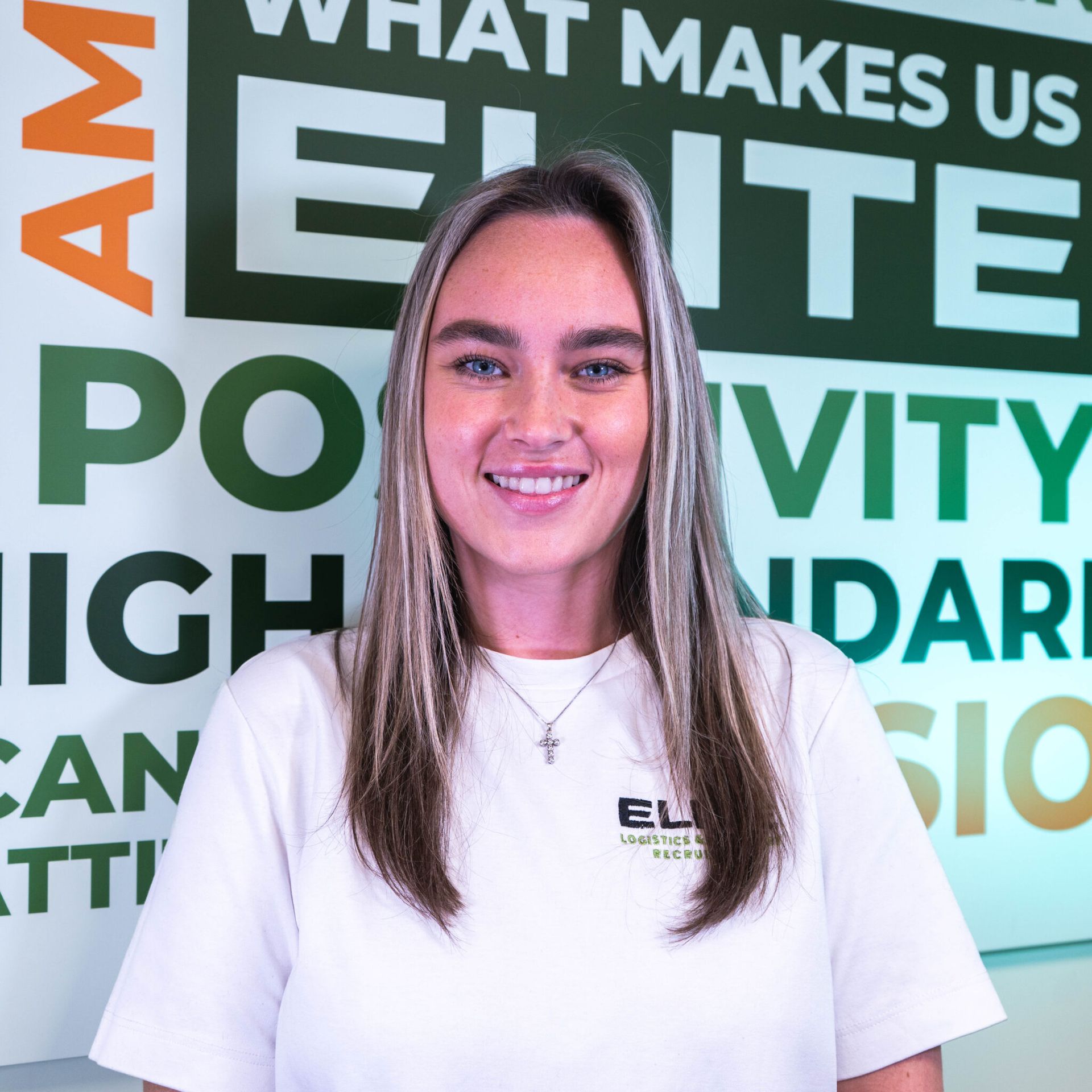 Woman with blonde hair smiles in front of a green wall with text, wearing a white shirt with an