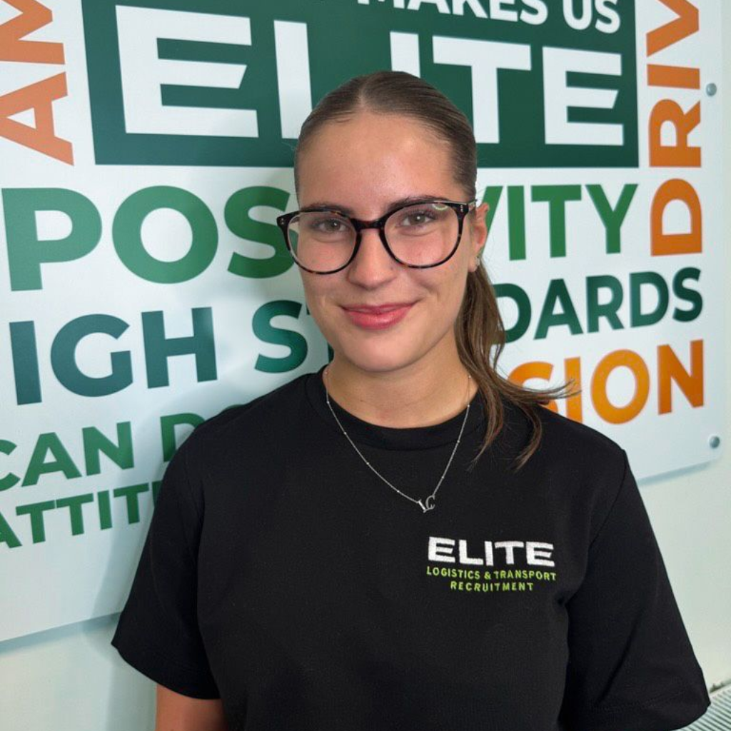 Woman with glasses smiling, wearing a black shirt in front of a green and white wall with text.