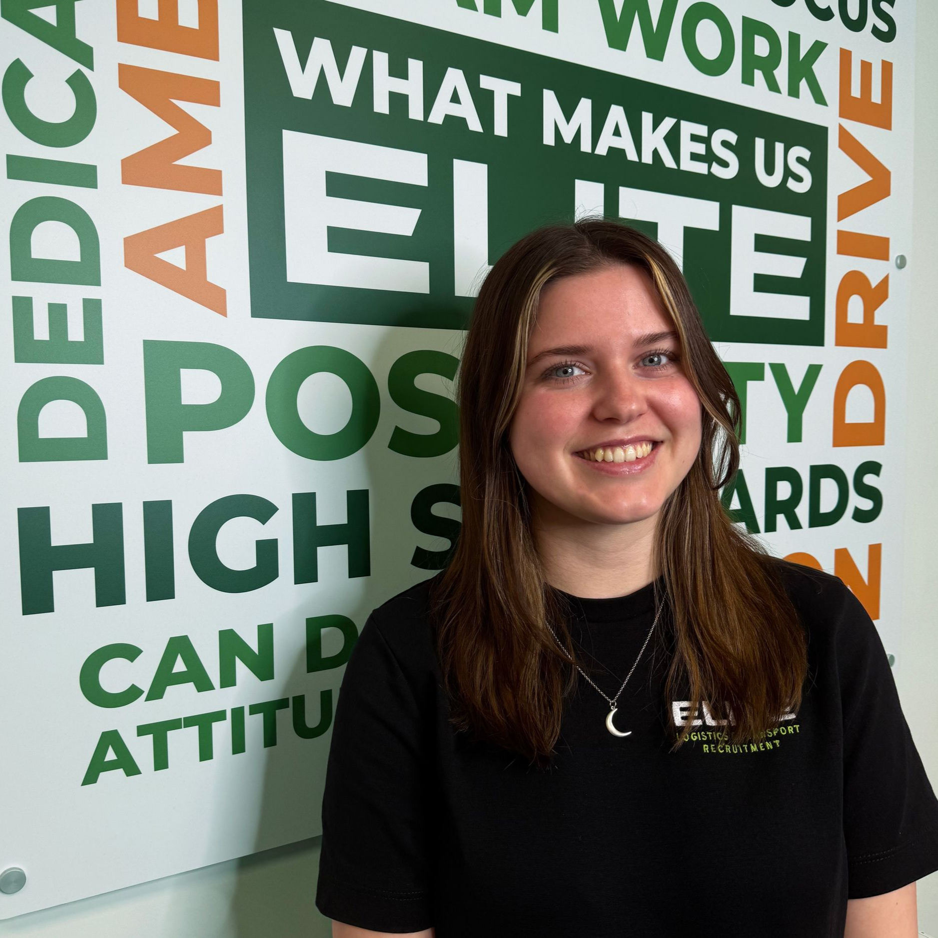 Woman with dark hair smiling, wearing a black shirt in front of a green and white wall with text.