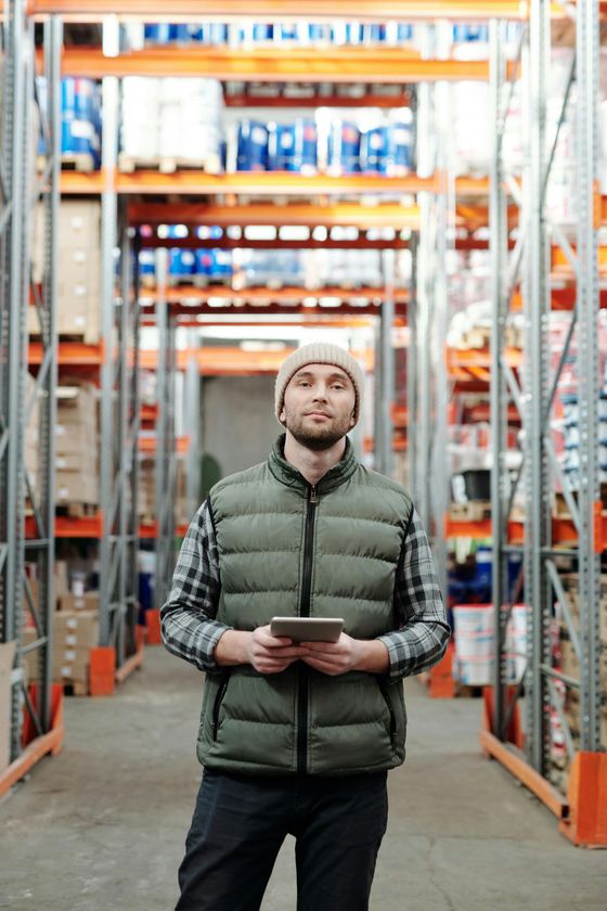 Man holding tablet, standing in warehouse aisle, wearing beanie and vest.